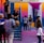 A vibrant outdoor basketball court filled with people of various ages engaged in different activities. Some are playing basketball while others are standing and chatting. The court is surrounded by brightly painted walls in shades of pink, orange, and blue. A basketball hoop is mounted on the back wall, adding to the sports theme. A few people are sitting on stairs or using phones, creating a relaxed and social atmosphere.