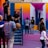 A vibrant outdoor basketball court filled with people of various ages engaged in different activities. Some are playing basketball while others are standing and chatting. The court is surrounded by brightly painted walls in shades of pink, orange, and blue. A basketball hoop is mounted on the back wall, adding to the sports theme. A few people are sitting on stairs or using phones, creating a relaxed and social atmosphere.