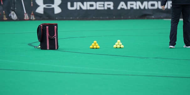 A sports field features a black and pink sports bag, two pyramids of yellow balls, and a portion of a person in dark athletic gear. The background displays a banner with the Under Armour logo.