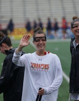 Several athletes in sports attire are on an outdoor sports field, interacting and smiling. One individual in the foreground waves with a friendly expression, while another stands beside her, also smiling. They wear protective goggles and gear associated with lacrosse. The background features indistinct figures in dark clothing against a backdrop of stadium seating.