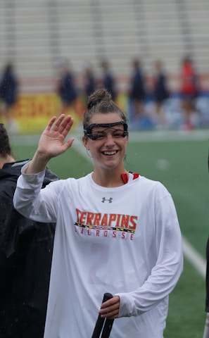 Several athletes in sports attire are on an outdoor sports field, interacting and smiling. One individual in the foreground waves with a friendly expression, while another stands beside her, also smiling. They wear protective goggles and gear associated with lacrosse. The background features indistinct figures in dark clothing against a backdrop of stadium seating.