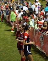 The image features a lively gathering of young soccer players and fans next to a field. Several female athletes in red and blue uniforms interact with an enthusiastic crowd standing behind a barrier. Some people in the crowd are holding objects, like pens and paper, possibly for autographs.