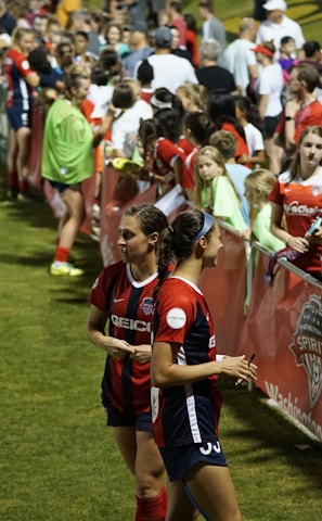 The image features a lively gathering of young soccer players and fans next to a field. Several female athletes in red and blue uniforms interact with an enthusiastic crowd standing behind a barrier. Some people in the crowd are holding objects, like pens and paper, possibly for autographs.