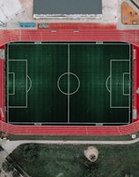 An aerial view of a large sports complex featuring a central green football field surrounded by a red running track. Adjacent to the track, there is a parking area, buildings, and pathways. Nearby, there are additional sports courts in different colors, including a blue swimming pool and orange basketball courts.