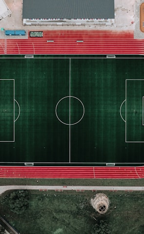 An aerial view of a large sports complex featuring a central green football field surrounded by a red running track. Adjacent to the track, there is a parking area, buildings, and pathways. Nearby, there are additional sports courts in different colors, including a blue swimming pool and orange basketball courts.