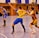 A dynamic indoor sports scene with players in motion on a court. Two athletes in blue and yellow uniforms are engaged in an intense handball game, with one player gripping the other's jersey. The background shows other players, several seated people, and advertisements on the walls.