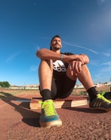 A person wearing a black athletic outfit is seated on a wooden platform in an outdoor sports field. The individual is relaxed, with arms resting on knees. The field is expansive with a clear blue sky and scattered clouds above. A brightly colored running shoe and athletic gear are visible, along with a water bottle and a blue backpack placed beside the person. Lampposts and residential buildings can be seen in the background.