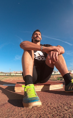 A person wearing a black athletic outfit is seated on a wooden platform in an outdoor sports field. The individual is relaxed, with arms resting on knees. The field is expansive with a clear blue sky and scattered clouds above. A brightly colored running shoe and athletic gear are visible, along with a water bottle and a blue backpack placed beside the person. Lampposts and residential buildings can be seen in the background.