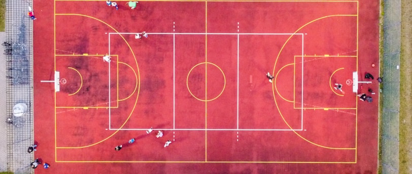 An aerial view of a sports court with a vibrant red surface and yellow lines marking different sections for basketball and soccer. Several people are present on the court, some engaged in activities, while others are standing along the perimeter. The court is enclosed by a metal fence and surrounded by grass on all sides.