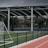 An outdoor sports facility featuring a running track and a soccer goal. The track is red, surrounded by a black metal fence, and the field is covered with artificial green turf. In the background, a basketball court is visible under a covered roof with metal supports. The sky is overcast, adding a moody atmosphere.