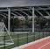An outdoor sports facility featuring a running track and a soccer goal. The track is red, surrounded by a black metal fence, and the field is covered with artificial green turf. In the background, a basketball court is visible under a covered roof with metal supports. The sky is overcast, adding a moody atmosphere.
