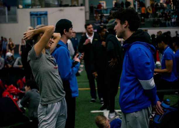 People engage in conversation and activities in an indoor sports facility. A woman in gray athletic wear stretches her arms above her head while talking to a man in a blue jacket. Other people are seen interacting in the background, some are seated on the ground, and the atmosphere is lively with many individuals gathered.