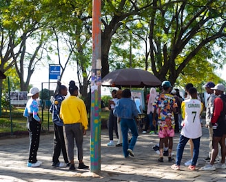 A group of people gathers in a park under leafy trees. They are interacting near a kiosk or stand surrounded by greenery and paved ground. Some individuals are wearing colorful clothing, and one person is wearing a number 7 sports jersey. There's a mixture of standing and walking activities happening in the scene.