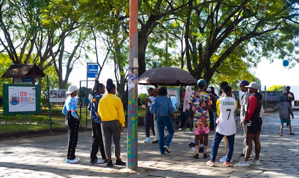 A group of people gathers in a park under leafy trees. They are interacting near a kiosk or stand surrounded by greenery and paved ground. Some individuals are wearing colorful clothing, and one person is wearing a number 7 sports jersey. There's a mixture of standing and walking activities happening in the scene.