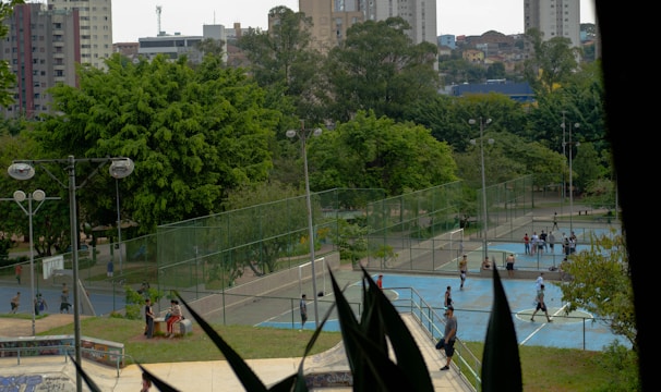 An outdoor recreational area in an urban setting featuring several sports courts and surrounded by lush green trees. People are engaged in various activities, including playing sports on the courts. In the background, there are tall buildings indicative of a cityscape.