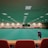 A large indoor sports facility featuring a green playing surface, similar to a bowling green. The ceiling is lined with numerous fluorescent lights, giving the space a bright yet serene atmosphere. Red chairs are arranged along the walls, and a scoreboard can be seen in the distance. A person is actively engaged in the game, bending forward as they release a ball, while inset a few spectators or participants are visible on the left side of the space.