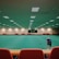 A large indoor sports facility featuring a green playing surface, similar to a bowling green. The ceiling is lined with numerous fluorescent lights, giving the space a bright yet serene atmosphere. Red chairs are arranged along the walls, and a scoreboard can be seen in the distance. A person is actively engaged in the game, bending forward as they release a ball, while inset a few spectators or participants are visible on the left side of the space.