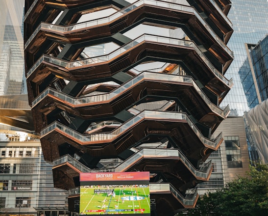 A large, spiral architectural structure with multiple levels stands prominently in an urban setting. In front of the structure, a large screen displays a sports broadcast, and several people are gathered around, some sitting and others standing. The scene is set outdoors, with towering modern buildings surrounding the area. The ground is paved with a geometric pattern of gray and white tiles.